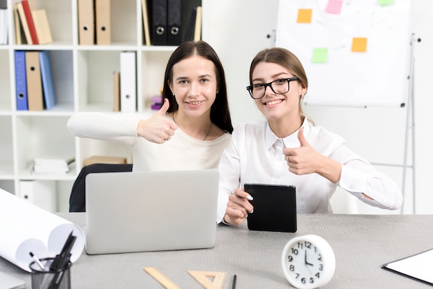 Free Photo smiling young businesswoman with laptop and digital tablet showing thumb up sign at workplace