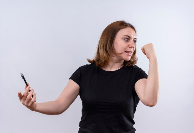 Smiling young casual woman holding mobile phone raising fist and looking at right side on isolated white space