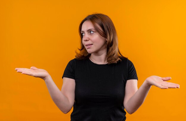 Smiling young casual woman looking at left side and showing empty hands on isolated orange space