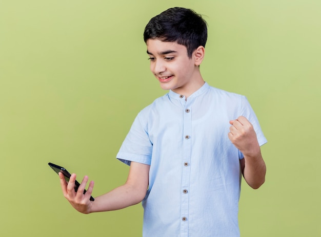 Free Photo smiling young caucasian boy holding and looking at mobile phone doing yes gesture isolated on olive green background