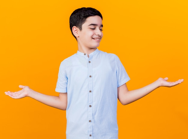 Free photo smiling young caucasian boy showing empty hands looking at them isolated on orange background