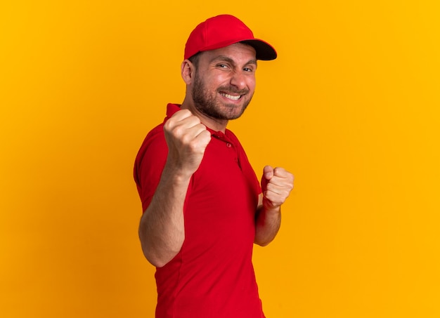 Smiling young caucasian delivery man in red uniform and cap standing in profile view looking at camera doing boxing gesture isolated on orange wall with copy space