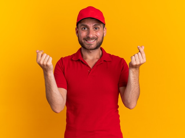 Free photo smiling young caucasian delivery man in uniform and cap doing money gesture