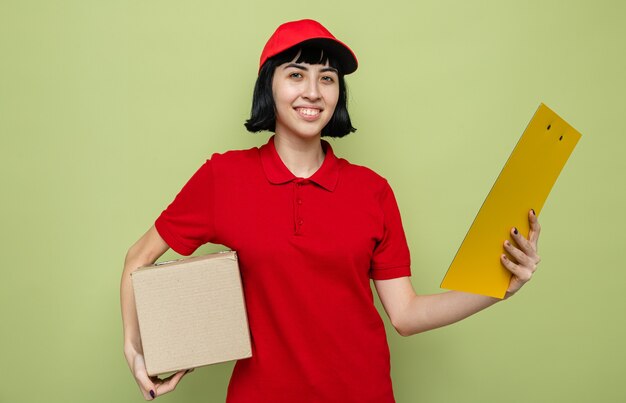 Smiling young caucasian delivery woman holding cardboard box and clipboard 