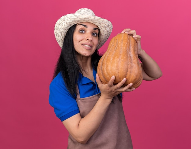 Free photo smiling young caucasian gardener girl wearing uniform and hat holding butternut pumpkin