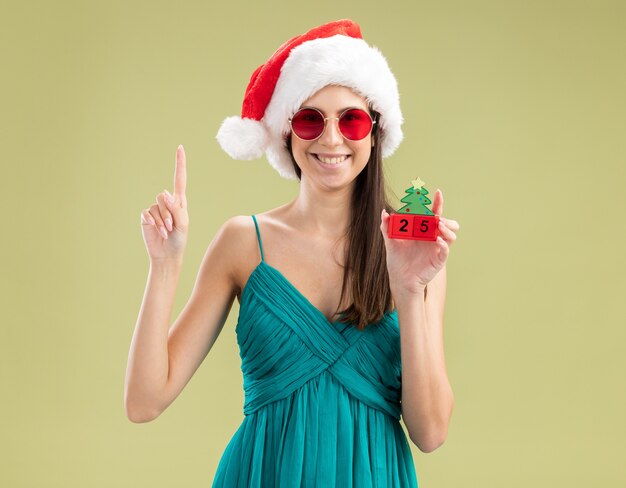 Smiling young caucasian girl in sun glasses with santa hat holding christmas tree ornament and pointing up 