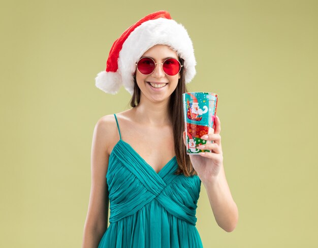 Smiling young caucasian girl in sun glasses with santa hat holding paper cup 