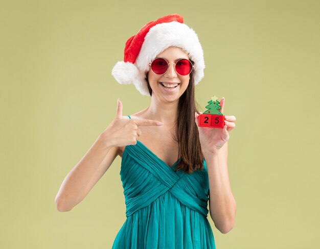 Smiling young caucasian girl in sun glasses with santa hat holding and pointing at christmas tree ornament 