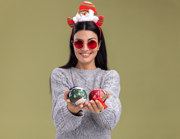 Smiling young caucasian girl wearing santa claus headband with glasses stretching out christmas baubles towards camera looking at camera isolated on olive green background with copy space