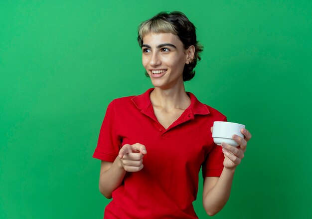 Smiling young caucasian girl with pixie haircut holding cup looking at side and pointing at camera isolated on green background with copy space