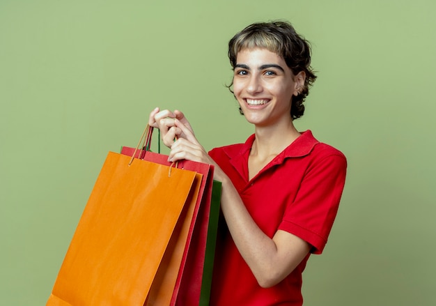 Smiling young caucasian girl with pixie haircut holding shopping bags isolated on olive green background