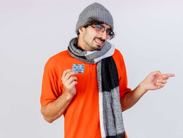 Free Photo smiling young caucasian ill man wearing glasses winter hat and scarf holding pack of medical capsules  pointing at side isolated on white wall