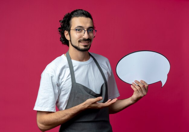 Smiling young caucasian male barber wearing glasses and wavy hair band in uniform holding chat bubble and pointing with hand at it isolated on crimson background