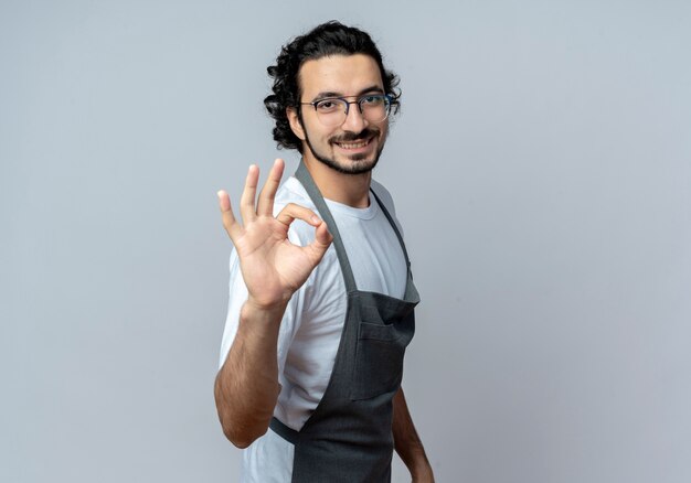 Free Photo smiling young caucasian male barber wearing glasses and wavy hair band in uniform standing in profile view doing ok sign