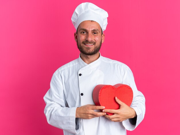 Smiling young caucasian male cook in chef uniform and cap holding heart shape 