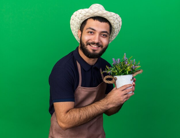 smiling young caucasian male gardener wearing gardening hat holding flowerpot isolated on green wall with copy space