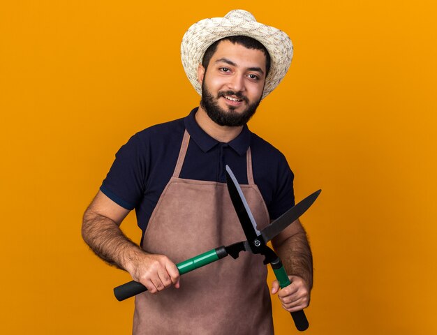 smiling young caucasian male gardener wearing gardening hat holding gardening scissors isolated on orange wall with copy space