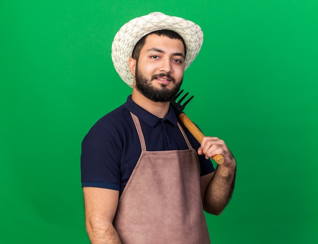 smiling young caucasian male gardener wearing gardening hat holding rake on shoulder isolated on green wall with copy space