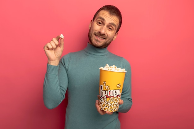 Smiling young caucasian man looking at camera holding bucket of popcorn and popcorn piece isolated on crimson background with copy space