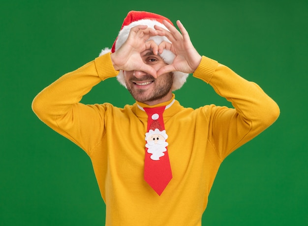 Free Photo smiling young caucasian man wearing christmas hat and tie looking at camera doing heart sign in front of eye isolated on green background