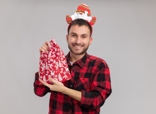 Smiling young caucasian man wearing christmas headband holding christmas sack looking at camera isolated on white background with copy space