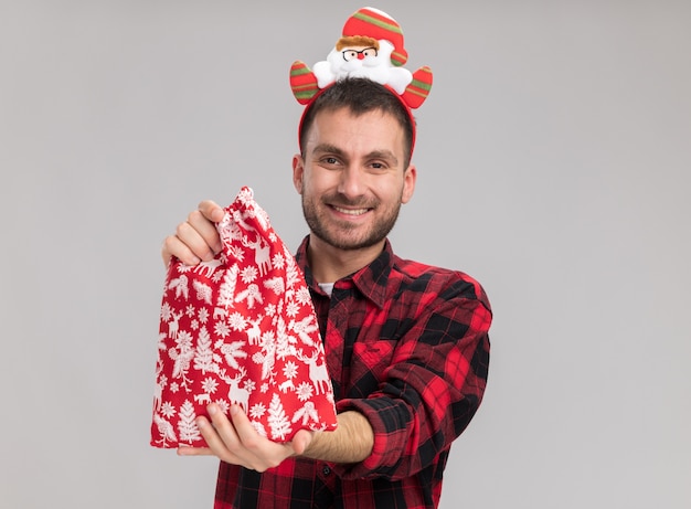 Smiling young caucasian man wearing christmas headband stretching out christmas sack towards camera looking at camera isolated on white background with copy space