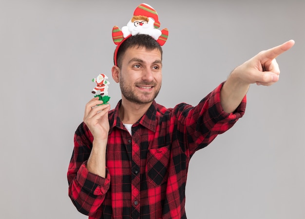 Free photo smiling young caucasian man wearing santa claus headband holding snowman christmas toy looking and pointing at side isolated on white background