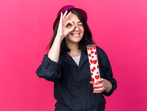 Free Photo smiling young caucasian party girl wearing party hat holding confetti cannon  doing look gesture isolated on pink wall