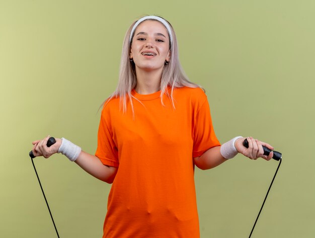 Smiling young caucasian sporty girl with braces wearing headband and wristbands holds jumping rope 