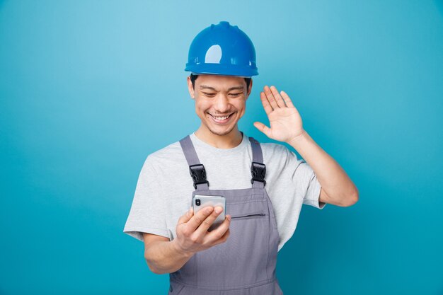 Smiling young construction worker wearing safety helmet and uniform holding and looking at mobile phone waving 