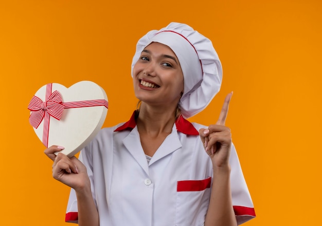 Smiling young cook female wearing chef uniform holding heart shape points finger to up box on isolated orange background