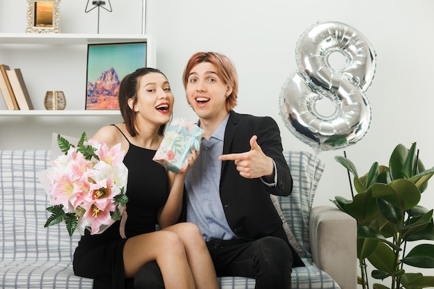 Smiling young couple on happy women day holding and points at bouquet and present sitting on sofa in living room