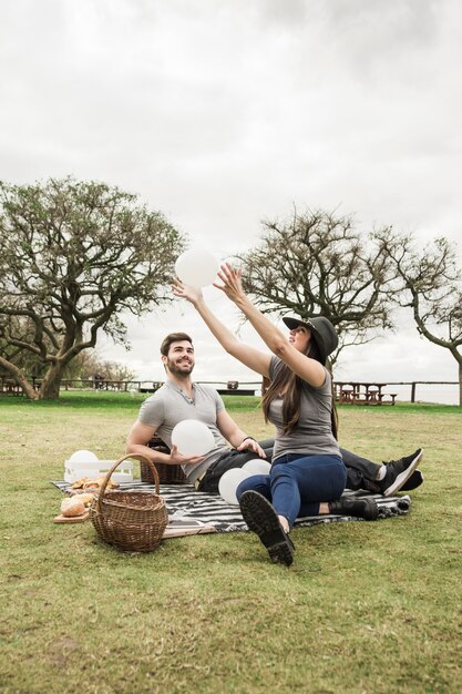 Smiling young couple playing with white balloons sitting in the park