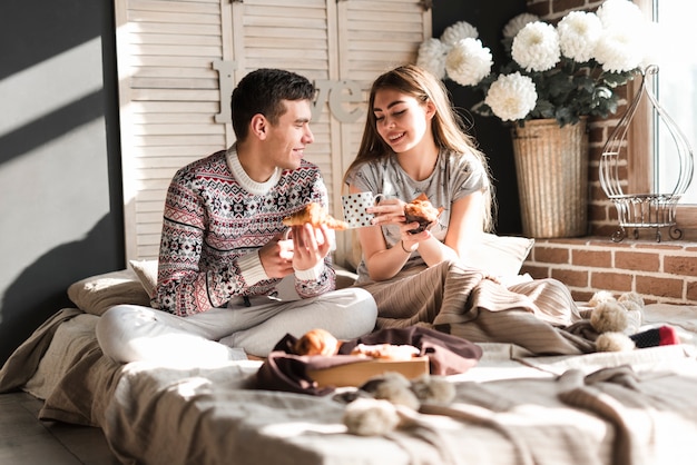 Smiling young couple sitting on bed holding croissant and cupcake in hand