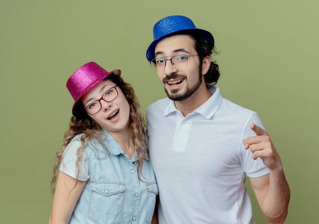 Free Photo smiling young couple wearing pink and blue hat guy showing you gesture isolated on olive green