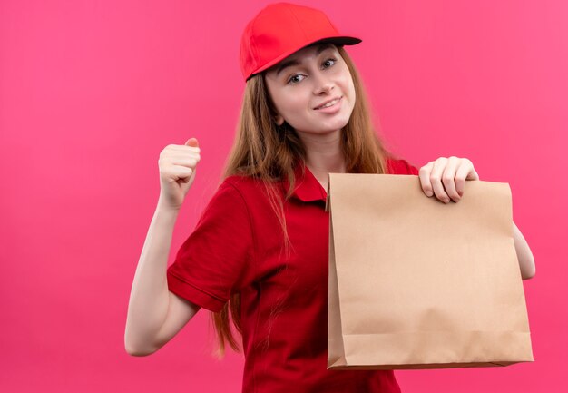 Smiling young delivery girl in red uniform holding paper bag with clenched fist on isolated pink wall