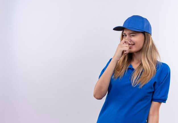 Smiling young delivery girl wearing blue uniform and cap closed nose isolated on white wall with copy space