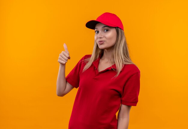 Smiling young delivery girl wearing red uniform and cap her thumb up isolated on orange wall