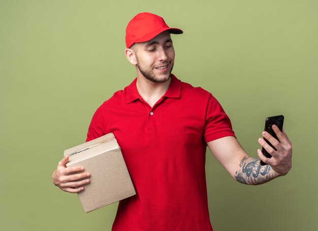 Smiling young delivery guy wearing uniform with cap holding box looking at phone in his hand 