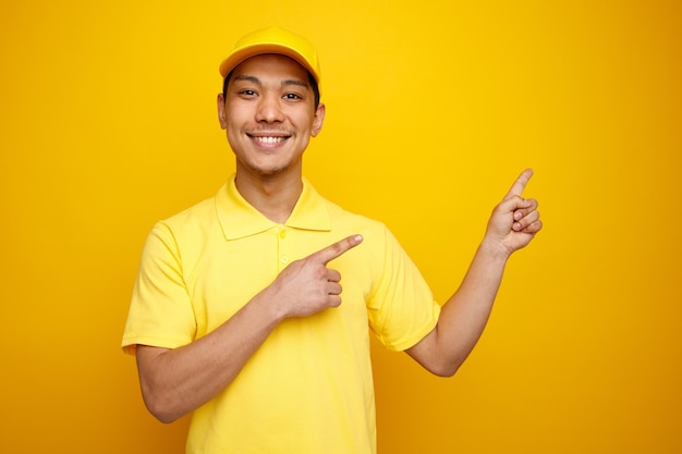 Smiling young delivery man wearing cap and uniform pointing up at corner 