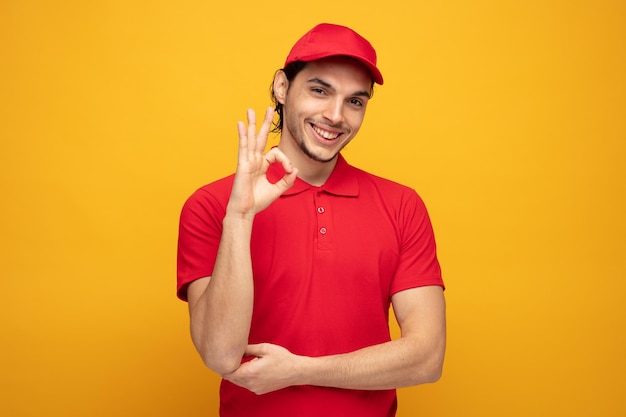 smiling young delivery man wearing uniform and cap looking at camera showing ok sign while keeping hand under elbow isolated on yellow background