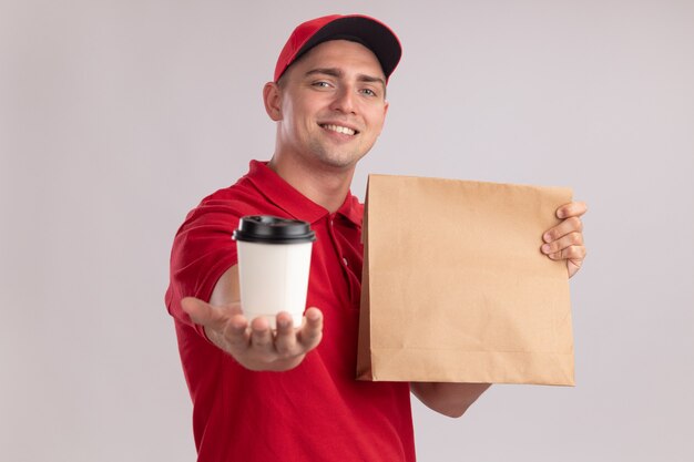 Smiling young delivery man wearing uniform with cap holding paper food package and holding out cup of coffee at front isolated on white wall