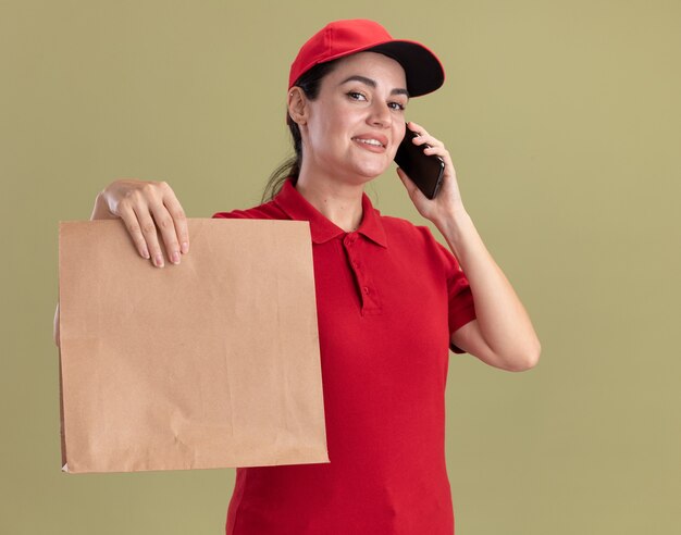 Smiling young delivery woman in uniform and cap talking on phone stretching out paper package towards camera 