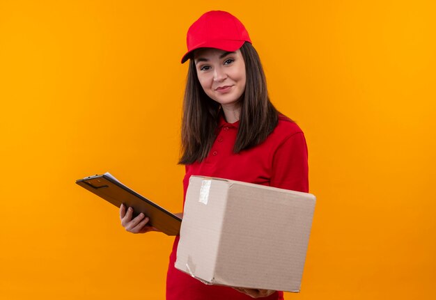 Smiling young delivery woman wearing red t-shirt in red cap holding a box and clipboard on isolated yellow wall