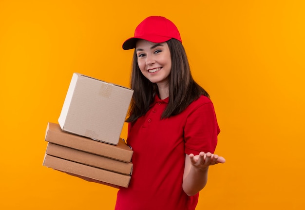 Smiling young delivery woman wearing red t-shirt in red cap holding a box and pizza box on isolated orange wall