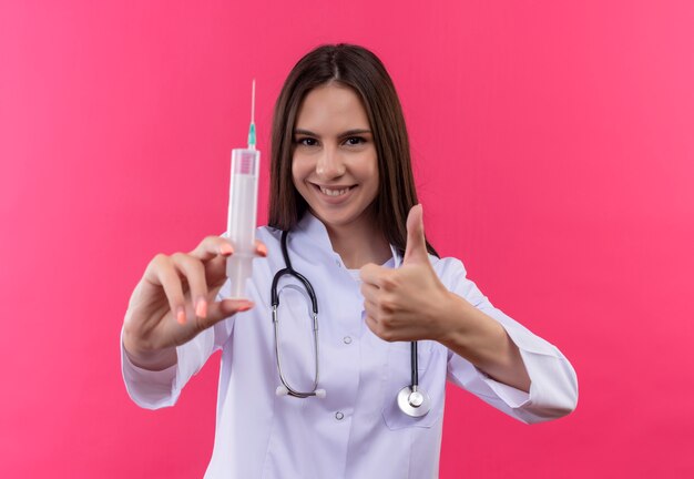 Smiling young doctor girl wearing stethoscope medical gown holding syringe her thumb up on isolated pink background