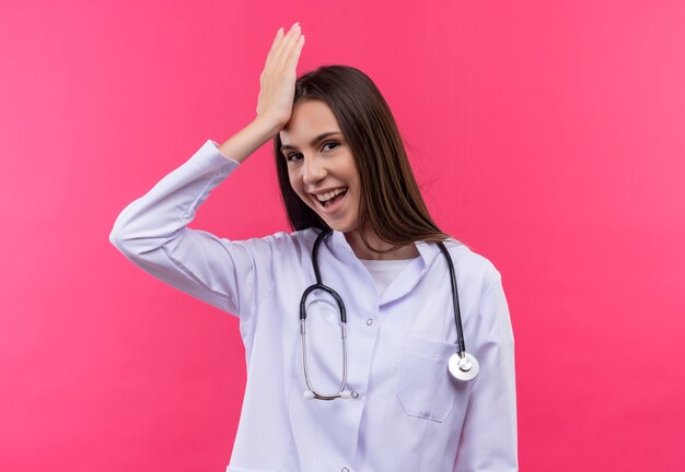 Smiling young doctor girl wearing stethoscope medical gown putting hand on nose on isolated pink background