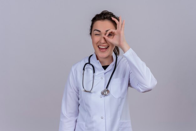 Smiling young doctor wearing medical gown wearing stethoscope shows okey gesture on white wall