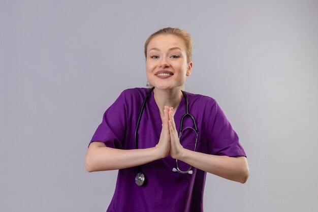 Smiling young doctor wearing purple medical gown and stethoscope shows pray gesture on isolated white wall