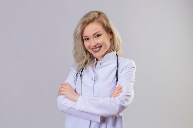 Smiling young doctor wearing stethoscope in medical gown crossing hands on white wall